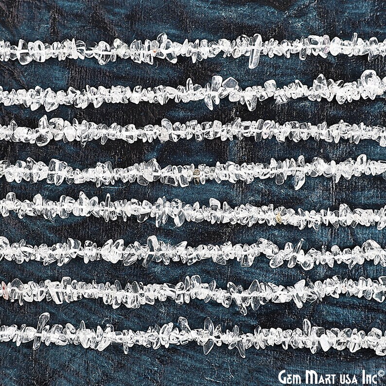 May include: A strand of clear quartz chip beads, arranged in rows on a dark blue background. The beads are small and irregular in shape, with a natural, unpolished finish.