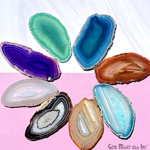 May include: Assortment of agate slices in various colors, including purple, blue, green, brown, white, and black. The slices are arranged in a circle on a white and pink surface. Gem Mart USA Inc. is printed in the bottom right corner.