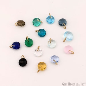 May include: A collection of round, faceted crystal pendants in various colors, including blue, green, pink, and clear, each with a small gold-colored loop for hanging. The pendants are scattered on a white surface.