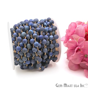 May include: A close-up of a spool of blue lapis lazuli beads strung on a gold chain. The beads are round and have a matte finish. The chain is delicate and has a gold finish.