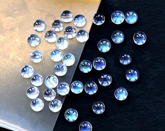 May include: A collection of round, iridescent moonstone cabochons in various sizes. The stones are scattered on a silver tray and a black surface. The stones have a blue and white color.