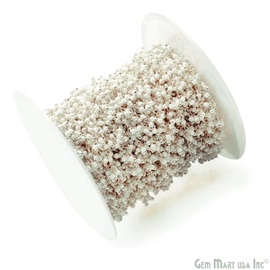 May include: A spool of silver chain with white beads. The chain is wound around a white spool.