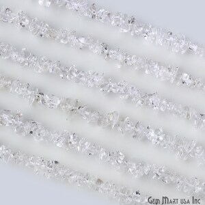 May include: A strand of clear quartz crystal chips, also known as rock crystal, arranged in parallel lines on a white background. The chips are irregular in shape and size, with some having a slightly frosted appearance.