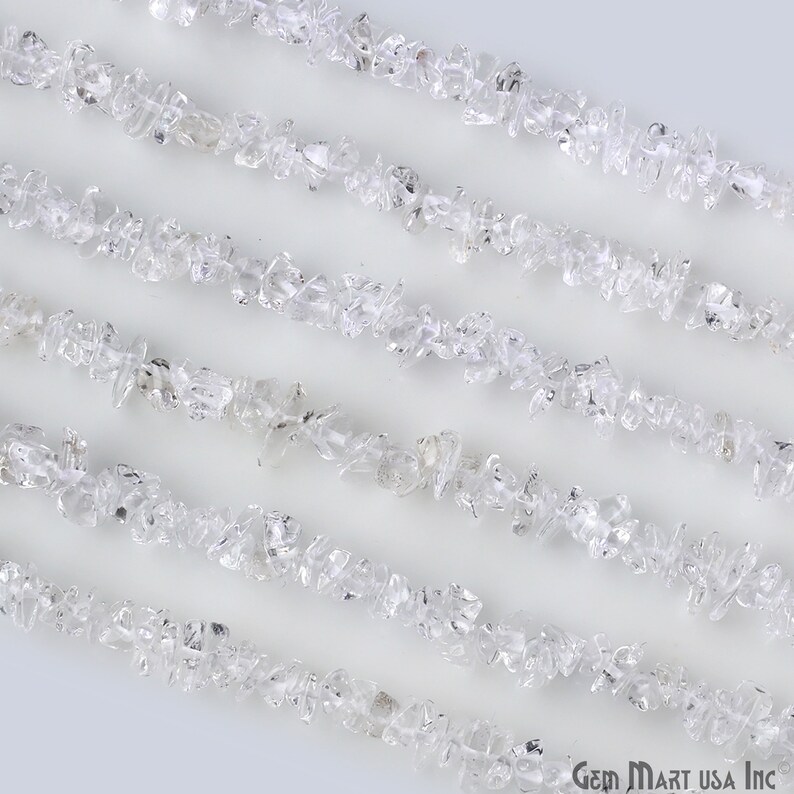 May include: A strand of clear quartz crystal chips, also known as rock crystal, arranged in parallel lines on a white background. The chips are irregular in shape and size, with some having a slightly frosted appearance.
