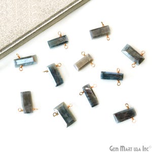 May include: A collection of rectangular, faceted labradorite gemstone beads with copper wire loops. The beads are various shades of gray and blue, scattered on a white surface. A gray, textured surface is in the upper left corner.