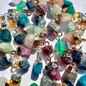 May include: A collection of raw gemstone pendants in various colors, including green, blue, pink, and gold. Each pendant features a unique, irregular shape and is capped with gold-colored metal and a small loop for a chain.