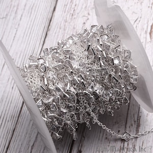 K&ouml;nnte beinhalten: Eine Spule silberner Kette mit klaren quadratischen Strasssteinen. Die Kette ist um eine wei&szlig;e Spule gewickelt.