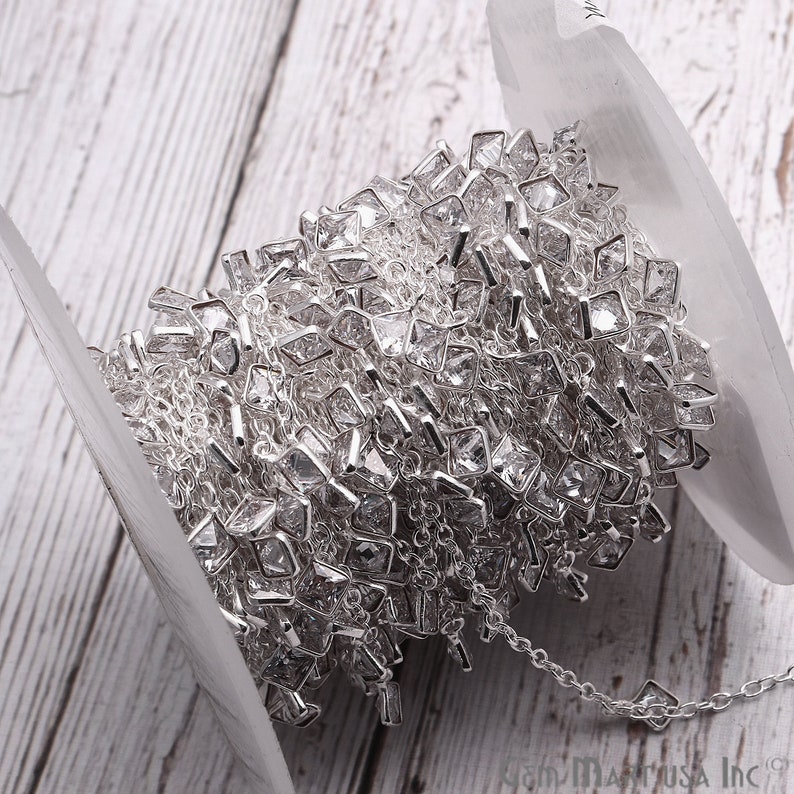 K&ouml;nnte beinhalten: Eine Spule silberner Kette mit klaren quadratischen Strasssteinen. Die Kette ist um eine wei&szlig;e Spule gewickelt.