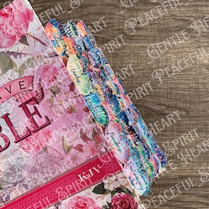 May include: A pink and white floral design Bible with a red spine and the words "PEACEFUL SPIRIT KJV" printed on the cover. The Bible has colorful tabs with the names of the books of the Bible printed on them.