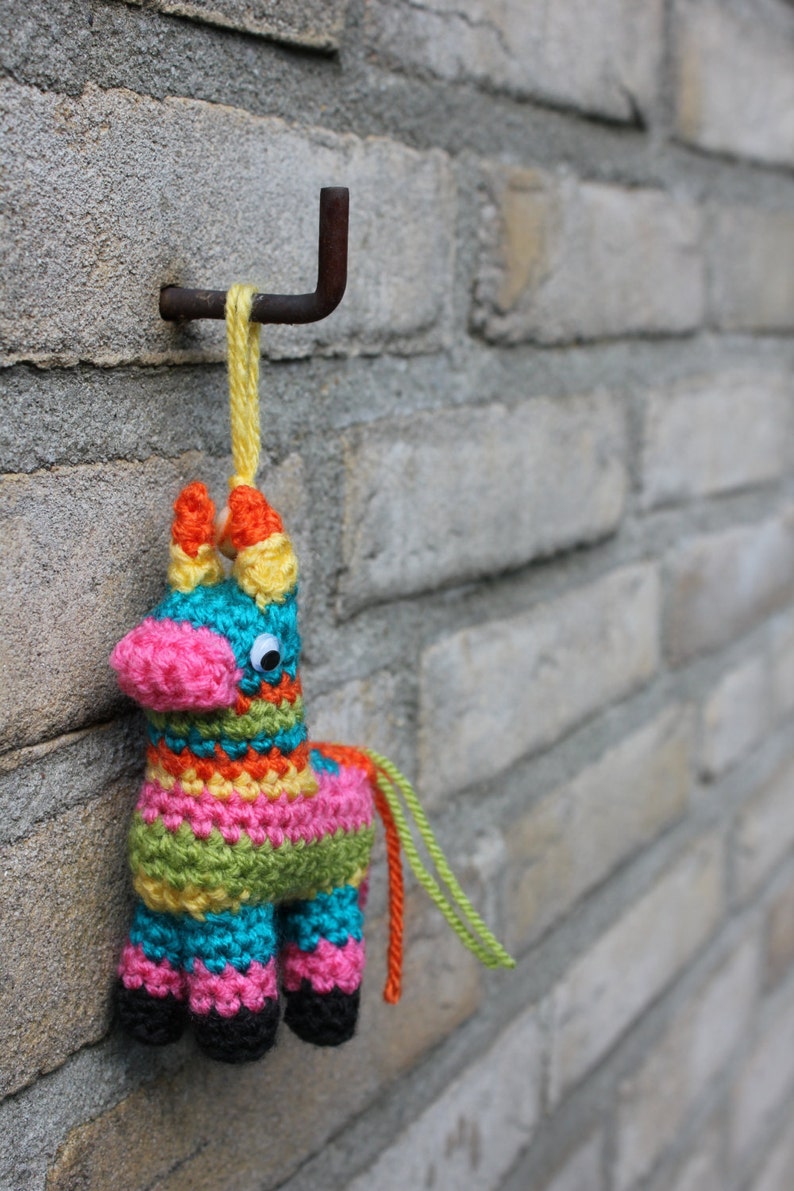 May include: A colorful crocheted pinata shaped like a donkey, hanging from a metal hook on a brick wall. The pinata is decorated with stripes of pink, yellow, green, blue, and orange yarn. It has a single googly eye and a long tail.