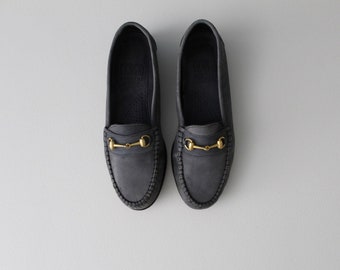 Könnte beinhalten: Ein Paar dunkelblaue Lederschuhe mit goldenen Pferdekopf-Akzenten. Die Schuhe haben einen genähten Rand und eine Ledersohle.