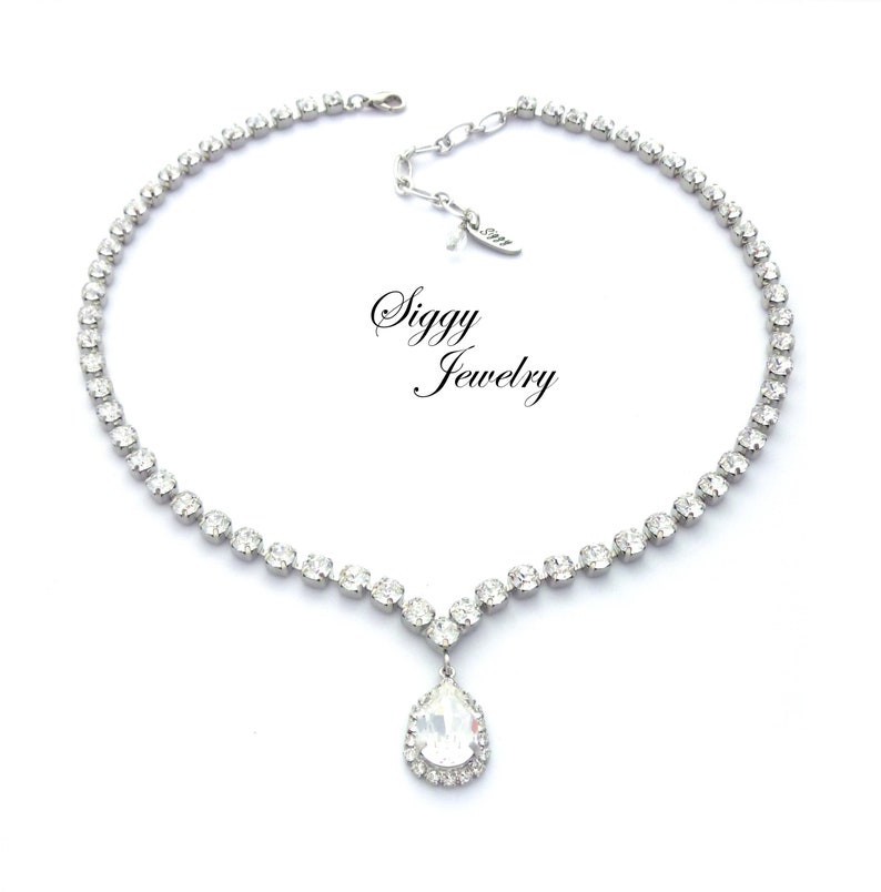 May include: A silver necklace with a teardrop-shaped pendant, both adorned with clear rhinestones. The necklace has a delicate chain and a clasp closure. The pendant is surrounded by a halo of smaller rhinestones. The necklace is labeled "Siggy Jewelry".