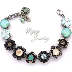 Puede incluir: Un brazalete de plata con un diseño floral. El brazalete presenta cristales de color azul claro, aguamarina y blanco, así como abalorios de flores de plata. El brazalete está acentuado con un abalorio de plata que dice "Siggy Jewelry".