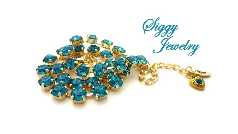 K&ouml;nnte beinhalten: Eine goldene Halskette mit einer Kette aus t&uuml;rkisblauen Kristallen und einem kleinen goldenen Herzanh&auml;nger mit einem t&uuml;rkisblauen Kristall. Die Halskette hat ein Etikett mit der Aufschrift "Siggy Jewelry".