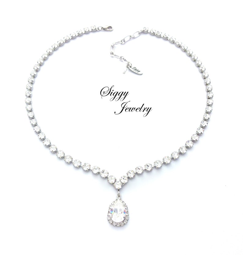 May include: A silver necklace with a teardrop-shaped pendant set with clear rhinestones. The necklace is made of small, clear rhinestones that are set in a silver chain. The pendant is also set with clear rhinestones and has a halo of smaller rhinestones around it. The necklace is labeled "Siggy Jewelry".