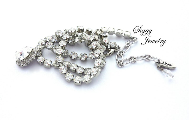 May include: A silver necklace with a teardrop-shaped crystal pendant and a chain of small crystals. The necklace has a silver clasp and a small tag with the word "Siggy" on it.
