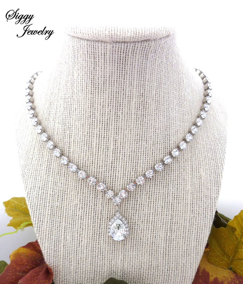 May include: A silver necklace with a teardrop-shaped pendant set with clear rhinestones. The necklace is on a white fabric-covered mannequin.
