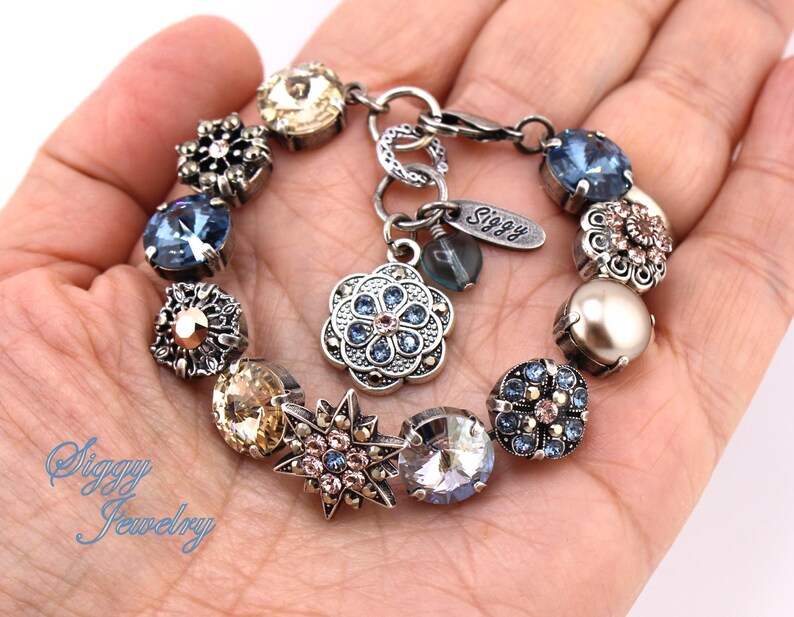 May include: A silver charm bracelet with a variety of blue, gold, and pink charms. The charms include flowers, stars, and crystals. The bracelet has a silver clasp and a tag that says "Siggy".