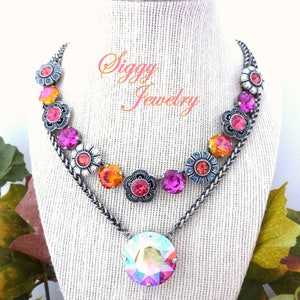 May include: A multi-layered necklace with a large iridescent crystal pendant. The necklace features a chain with a row of silver flower charms with pink, orange, and blue crystals.