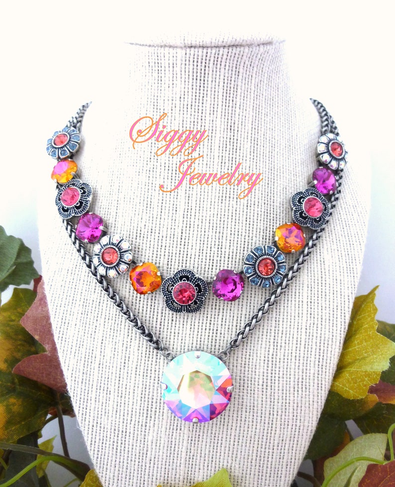 May include: A multi-layered necklace with a large iridescent crystal pendant. The necklace features a chain with a row of silver flower charms with pink, orange, and blue crystals.