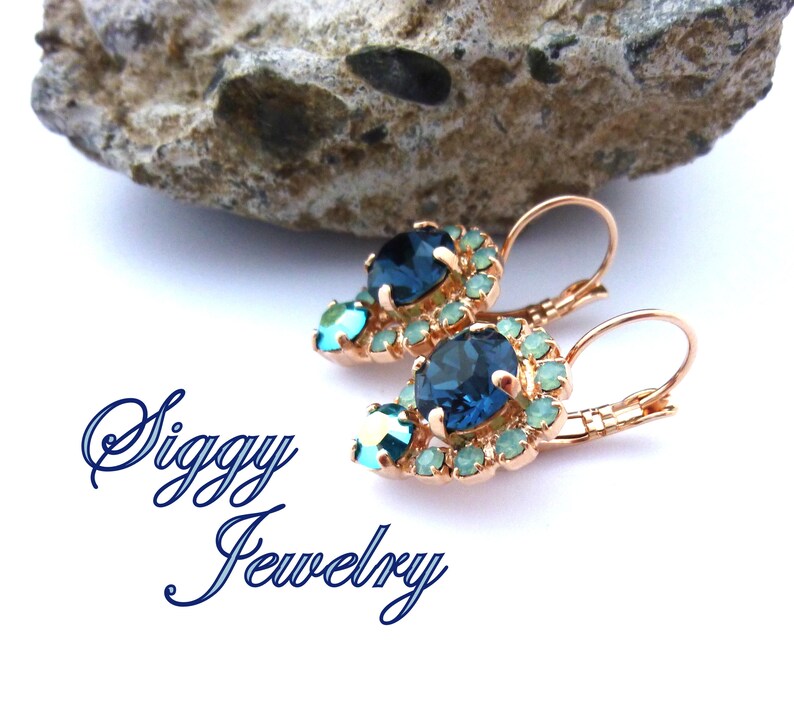 Pu&ograve; includere: Un paio di orecchini pendenti con un cristallo blu a forma di lacrima circondato da cristalli pi&ugrave; piccoli di colore blu chiaro. Gli orecchini sono in metallo dorato rosa e hanno una chiusura a gancio.