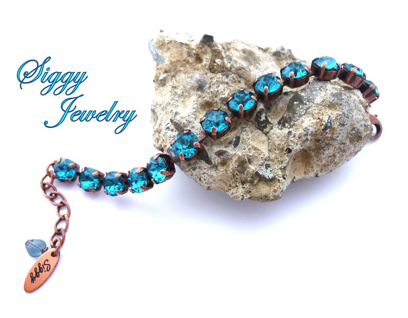 K&ouml;nnte beinhalten: Ein Kupferarmband mit t&uuml;rkisblauen Kristallen. Das Armband liegt auf einem Stein mit dem Schriftzug "Siggy Jewelry" im Hintergrund.