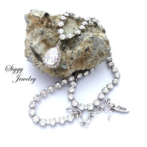 May include: A silver necklace with a teardrop-shaped crystal pendant. The necklace is made of small, clear rhinestones and has a silver clasp. The necklace is on a rock with the text "Siggy Jewelry" on the left side of the image.