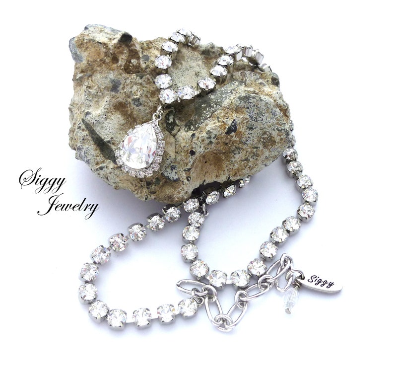 May include: A silver necklace with a teardrop-shaped crystal pendant. The necklace is made of small, clear rhinestones and has a silver clasp. The necklace is on a rock with the text "Siggy Jewelry" on the left side of the image.