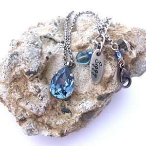 Op de afbeelding: Een zilveren ketting met een traanvormige blauwe kristallen hanger. De ketting heeft een zilveren ketting en een zilveren bedel met de gravure "Siggy".