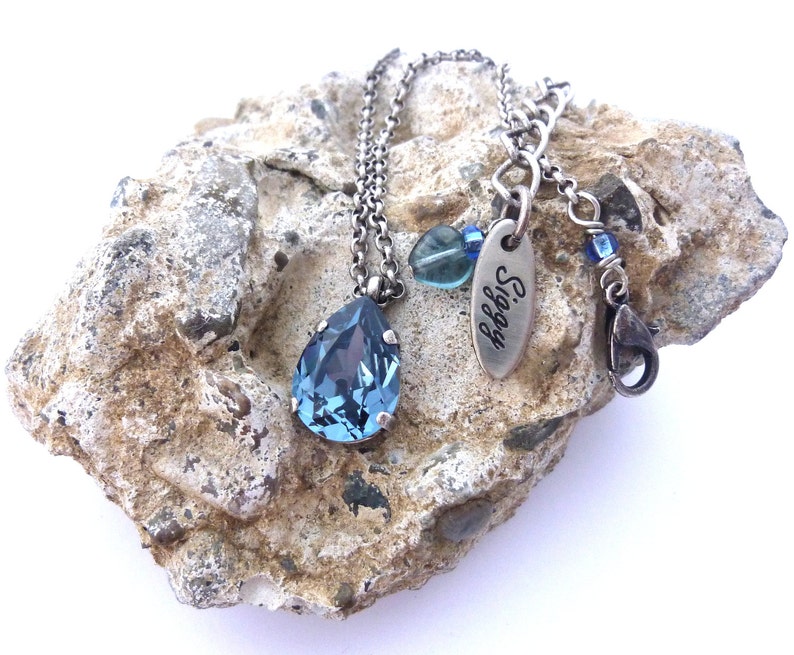 Op de afbeelding: Een zilveren ketting met een traanvormige blauwe kristallen hanger. De ketting heeft een zilveren ketting en een zilveren bedel met de gravure "Siggy".
