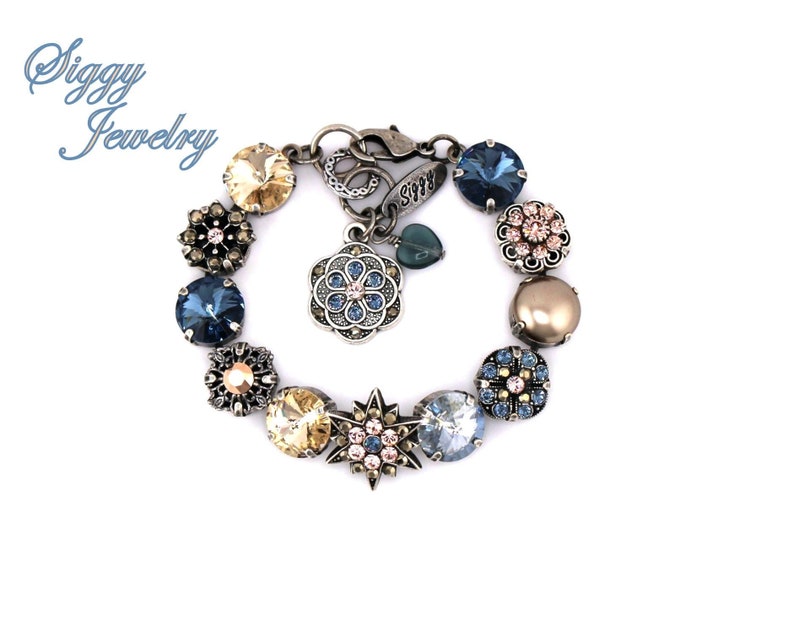 May include: A silver charm bracelet with a variety of blue, gold, and pink crystal charms. The bracelet features a flower charm with a blue crystal center and a star charm with a pink crystal center. The bracelet also includes a charm with the word "Siggy" on it.