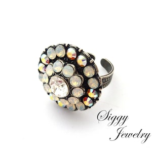 May include: A silver ring with a large, round, clear crystal surrounded by smaller, iridescent crystals. The ring is set on a white background.