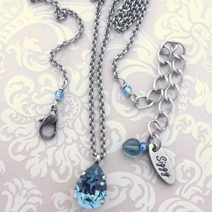 Op de afbeelding: Een zilveren ketting met een traanvormige blauwe kristallen hanger. De ketting heeft een karabijn sluiting en een klein bedeltje met het woord "Siggy" erop gegraveerd.