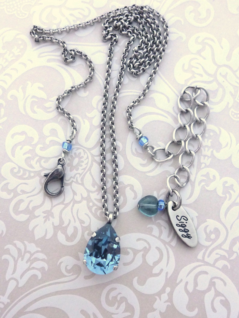 Op de afbeelding: Een zilveren ketting met een traanvormige blauwe kristallen hanger. De ketting heeft een karabijn sluiting en een klein bedeltje met het woord "Siggy" erop gegraveerd.