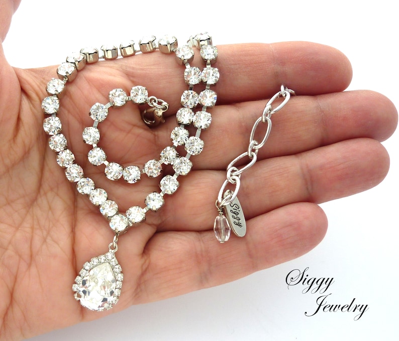 May include: A silver necklace with a teardrop-shaped crystal pendant and a chain of small round crystals. The necklace has a silver clasp and a small tag that says "Siggy".