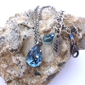 Op de afbeelding: Een zilveren ketting met een traanvormige blauwe kristallen hanger. De ketting heeft een zilveren tag met het woord "Siggy" erop gegraveerd.