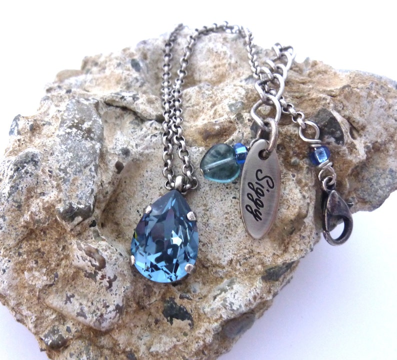 Op de afbeelding: Een zilveren ketting met een traanvormige blauwe kristallen hanger. De ketting heeft een zilveren tag met het woord "Siggy" erop gegraveerd.