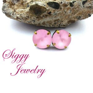 Pode incluir: Um par de brincos de press&atilde;o rosa com detalhes dourados. Os brincos est&atilde;o sobre um fundo branco com uma rocha atr&aacute;s. O texto "Siggy Jewelry" &eacute; vis&iacute;vel abaixo dos brincos.