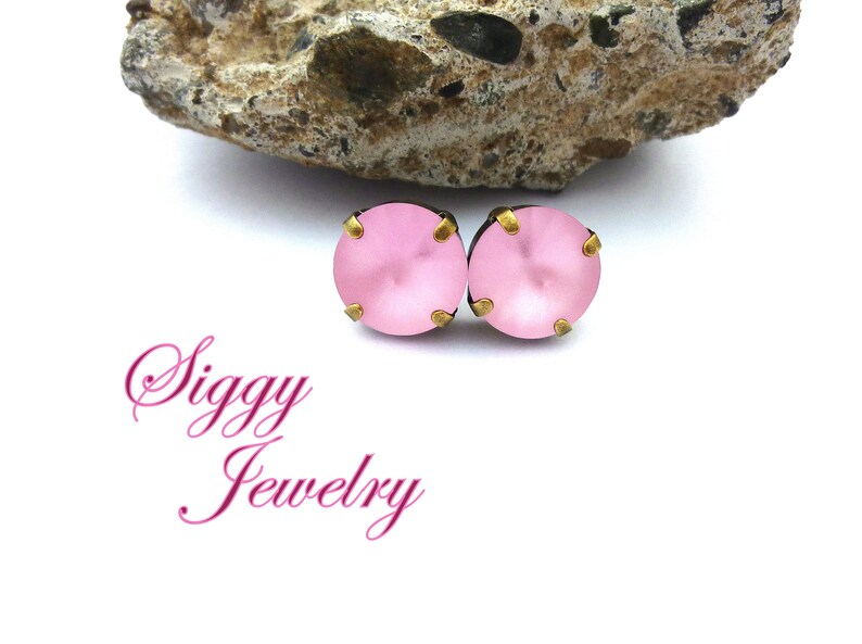 Pode incluir: Um par de brincos de press&atilde;o rosa com detalhes dourados. Os brincos est&atilde;o sobre um fundo branco com uma rocha atr&aacute;s. O texto "Siggy Jewelry" &eacute; vis&iacute;vel abaixo dos brincos.