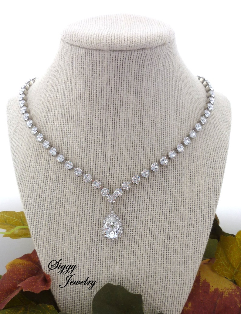 May include: A silver necklace with a teardrop-shaped pendant, both adorned with clear rhinestones. The necklace is displayed on a fabric-covered mannequin.