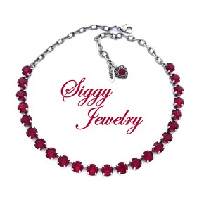 May include: A silver chain necklace with a red crystal stone design. The necklace has a small silver charm with a floral design. The text "Siggy Jewelry" is visible in the image.