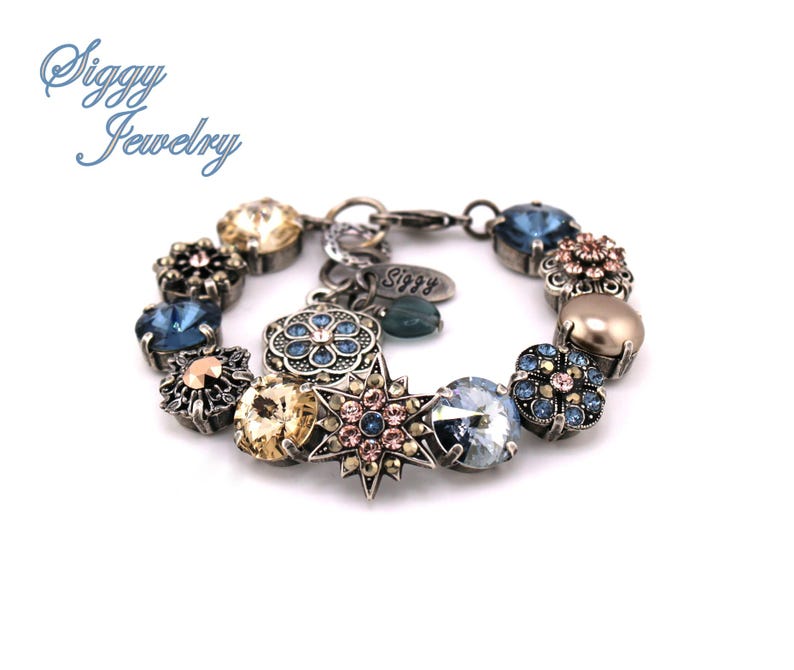 May include: A silver charm bracelet with a variety of blue, pink, and gold crystal charms. The charms include flowers, stars, and a round charm with a floral design. The bracelet has a silver clasp and a small tag that says "Siggy".