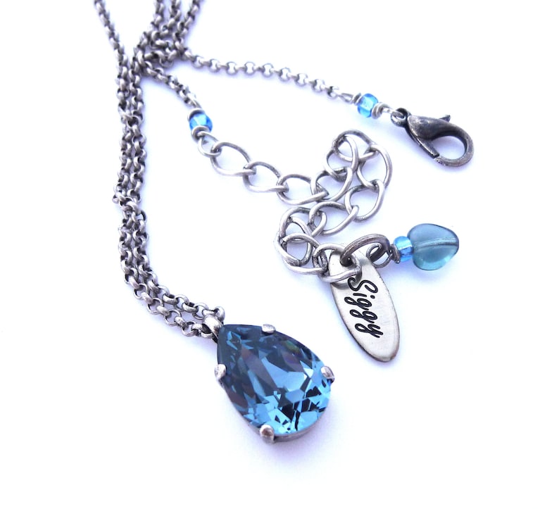 Op de afbeelding: Een zilveren ketting met een traanvormige blauwe kristallen hanger. De ketting heeft een zilveren bedel met het woord "Siggy" erop gegraveerd.