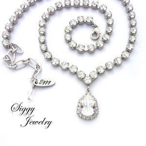 May include: A silver necklace with a teardrop-shaped crystal pendant. The necklace is made of small, clear crystals and has a delicate chain. The pendant is surrounded by a halo of smaller crystals. The necklace is labeled "Siggy Jewelry".