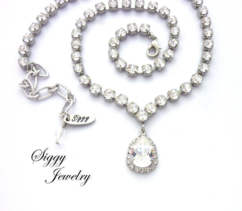 May include: A silver necklace with a teardrop-shaped crystal pendant. The necklace is made of small, clear crystals and has a delicate chain. The pendant is surrounded by a halo of smaller crystals. The necklace is labeled "Siggy Jewelry".