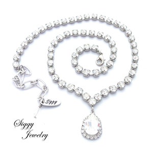 May include: A silver necklace with a teardrop-shaped crystal pendant. The necklace is made of small, clear rhinestones and has a lobster clasp closure. The pendant is surrounded by a halo of smaller rhinestones. The necklace is labeled "Siggy Jewelry".
