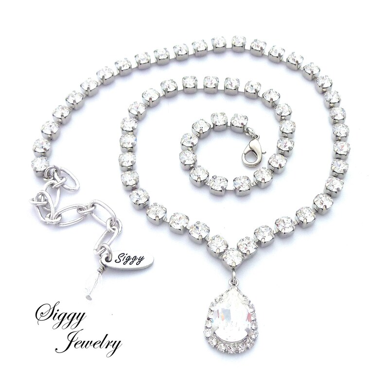 May include: A silver necklace with a teardrop-shaped crystal pendant. The necklace is made of small, clear rhinestones and has a lobster clasp closure. The pendant is surrounded by a halo of smaller rhinestones. The necklace is labeled "Siggy Jewelry".