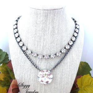 May include: A silver chain necklace with a large clear crystal pendant and a smaller row of clear crystals around the neck. The necklace is displayed on a white fabric mannequin bust. The text "Siggy Jewelry" is visible at the bottom of the image.