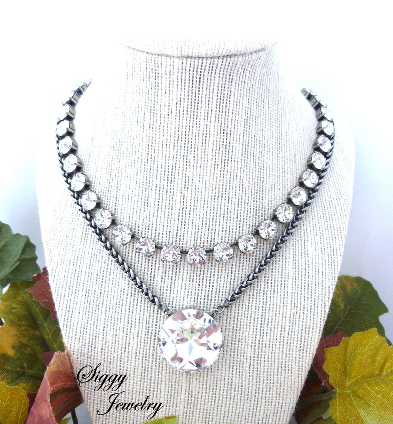 May include: A silver chain necklace with a large clear crystal pendant and a smaller row of clear crystals around the neck. The necklace is displayed on a white fabric mannequin bust. The text "Siggy Jewelry" is visible at the bottom of the image.