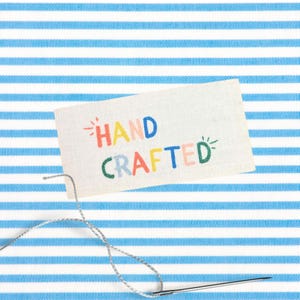 May include: A white rectangular tag with the words "HAND CRAFTED" in colorful embroidered letters. A needle and thread are positioned below the tag, against a blue and white striped background.